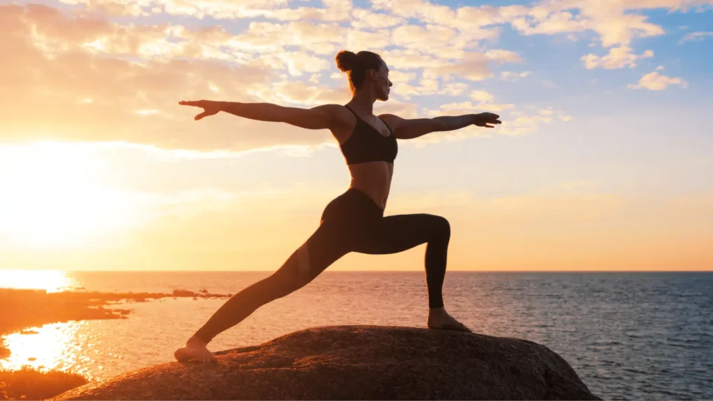Person practicing Warrior One yoga pose on a rock by the ocean at sunset