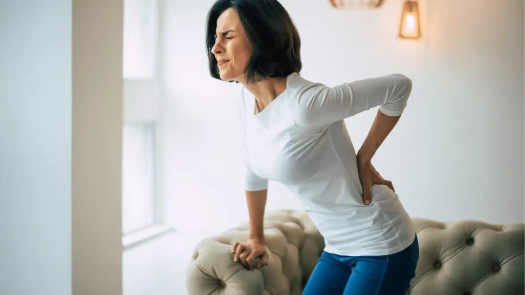 Woman holding her lower back in severe pain while standing in a living room