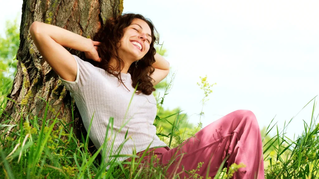 Smiling woman leaning back against a tree trunk in a sunny, grassy field